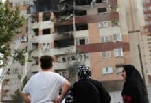 The image of a three people stand looking at a damaged residential building in Tehran, Iran, with blackened windows and visible structural destruction caused by an airstrike. A firefighter’s ladder reaches up to the upper floors.