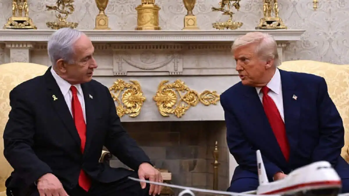 US President Donald Trump (right) and Israeli Prime Minister Benjamin Netanyahu (left) sit facing each other in a formal setting, likely the Oval Office. Both are wearing dark suits with red ties. A model airplane is placed on the table between them. Behind them is an ornate fireplace decorated with gold vases and intricate embellishments, highlighting the room's grandeur.