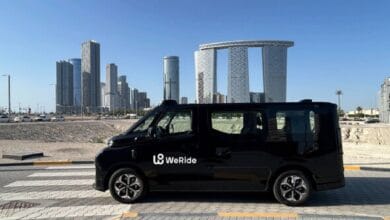 The image of a black WeRide autonomous vehicle is parked on a road with modern high-rise buildings and a clear blue sky in the background.