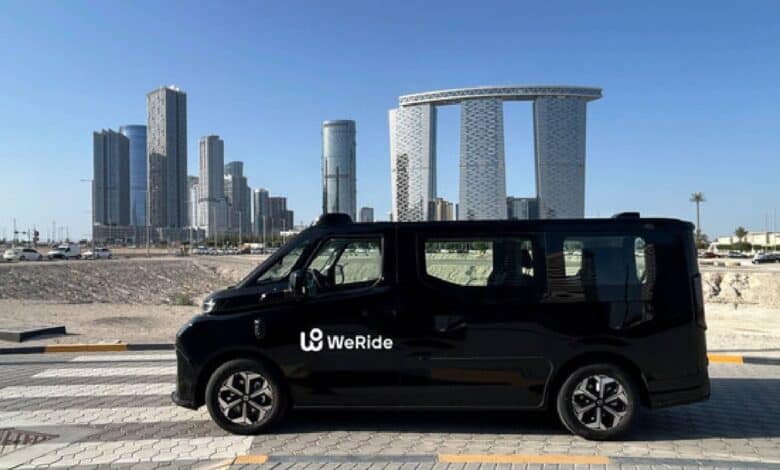The image of a black WeRide autonomous vehicle is parked on a road with modern high-rise buildings and a clear blue sky in the background.