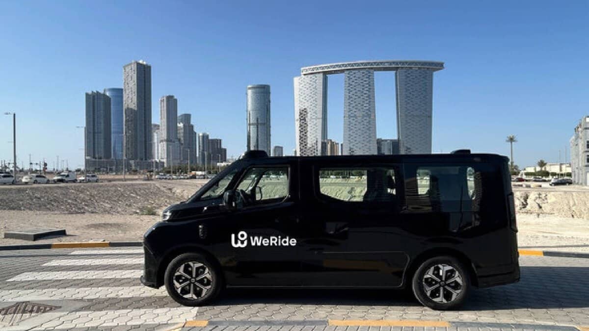 The image of a black WeRide autonomous vehicle is parked on a road with modern high-rise buildings and a clear blue sky in the background.