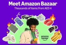 A promotional image for Amazon Bazaar featuring a smiling person with curly hair holding a smartphone, surrounded by various products including a phone case, sunglasses, slippers, a belt, a shirt, a travel pillow, and a wallet, with flames and heart emojis. The background is purple, and the text reads "Meet Amazon Bazaar Thousands of items from Dirham 4" with the Amazon Bazaar logo.