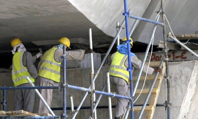 The image of construction workers wearing safety gear and helmets work on scaffolding under a concrete structure.