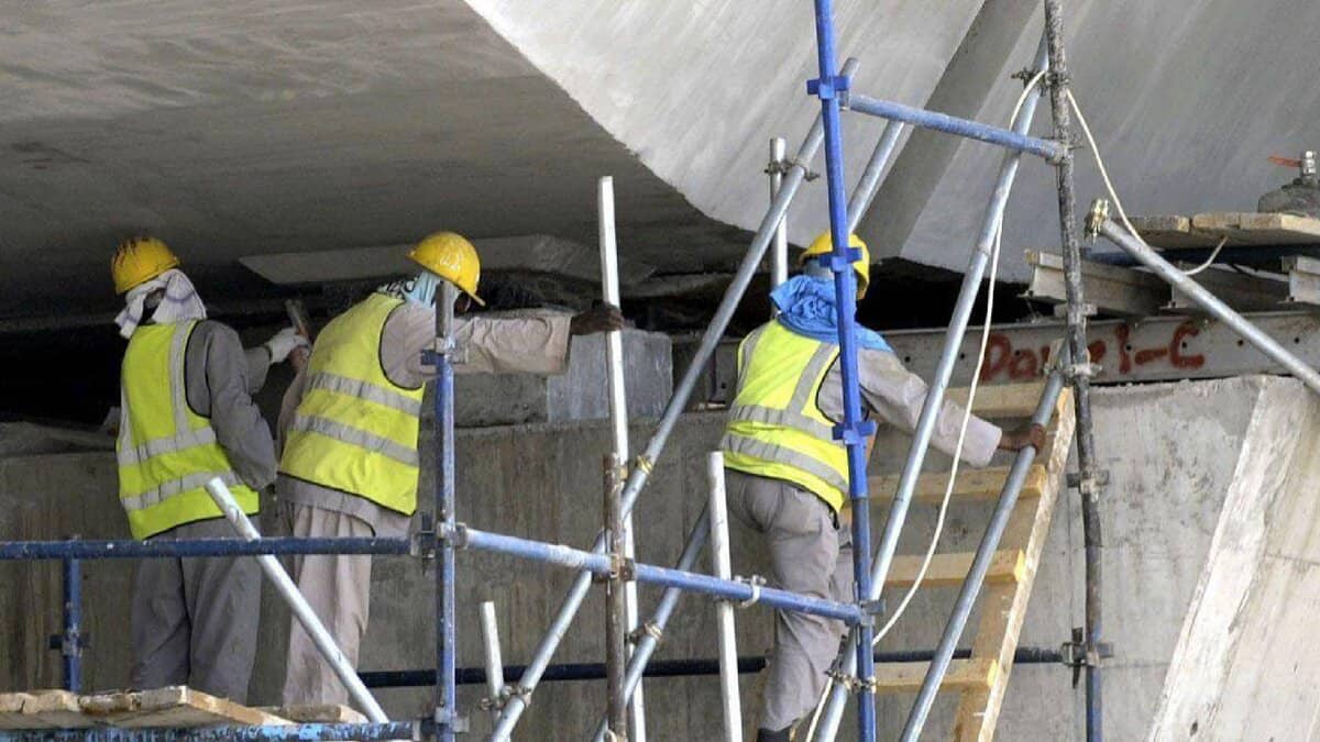 The image of construction workers wearing safety gear and helmets work on scaffolding under a concrete structure.