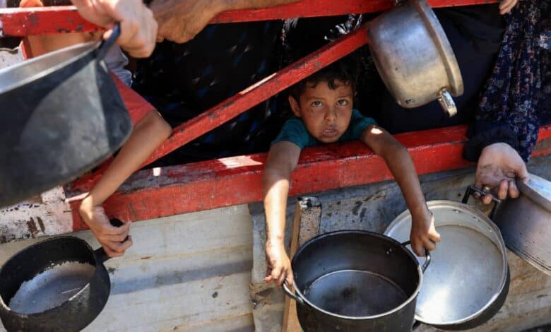 A young child with a somber expression peers out from a weathered red-framed structure in Gaza, clutching a metal pot. Surrounding hands of adults, also holding pots and pans, are visible.