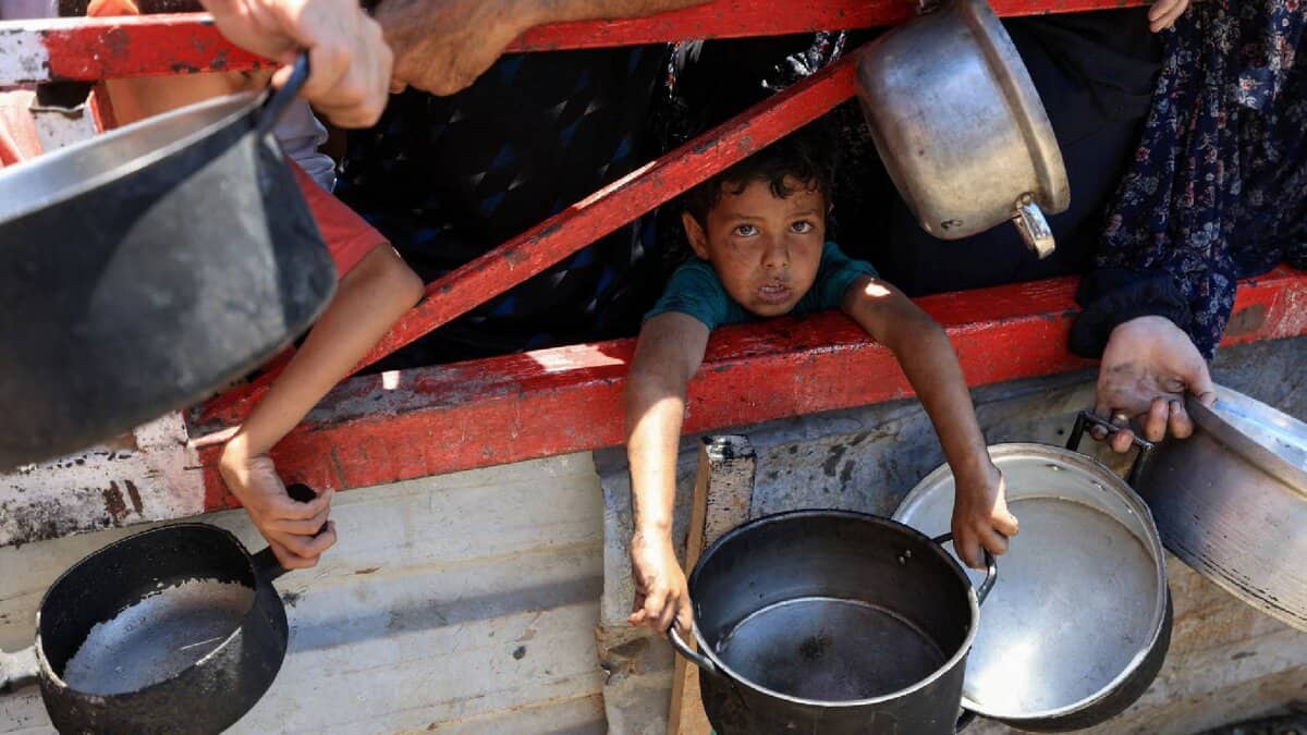 A young child with a somber expression peers out from a weathered red-framed structure in Gaza, clutching a metal pot. Surrounding hands of adults, also holding pots and pans, are visible.