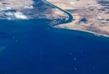 Aerial view of cargo ships queued in the Gulf of Suez near the entrance to the Suez Canal, with desert landscapes and scattered clouds above.
