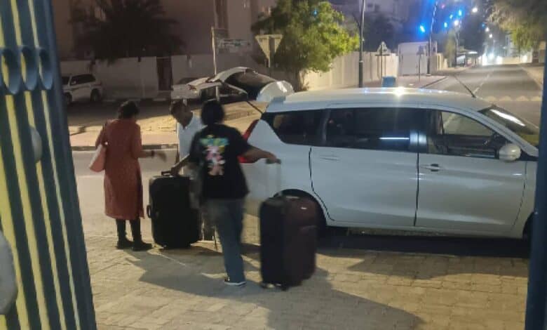 Two Indian women with suitcases are assisted by a man while preparing to board a white vehicle on a quiet street in Doha at night. A well-lit urban background with palm trees and modern buildings is visible.