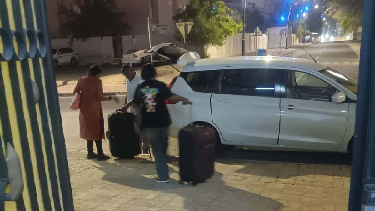 Two Indian women with suitcases are assisted by a man while preparing to board a white vehicle on a quiet street in Doha at night. A well-lit urban background with palm trees and modern buildings is visible.
