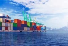 Stacked shipping containers at a busy seaport with cranes by the water.