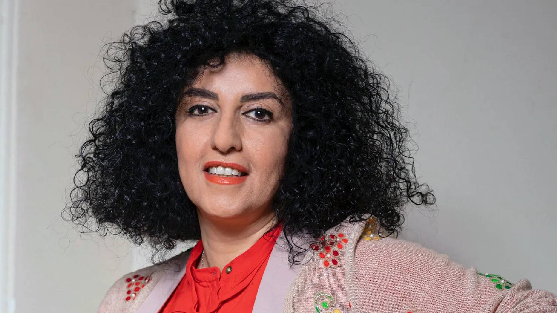 Narges Mohammadi, Iranian human rights activist and Nobel Peace Prize laureate, poses in a red blouse and embroidered jacket, smiling slightly with her curly black hair framing her face.