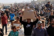 A large crowd of Palestinians, including men, women, and children, gathers near an aid distribution site in Rafah, southern Gaza Strip, with some holding boxes and bags of supplies.