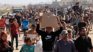 A large crowd of Palestinians, including men, women, and children, gathers near an aid distribution site in Rafah, southern Gaza Strip, with some holding boxes and bags of supplies.