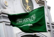 The national flag of Saudi Arabia waves in the wind in front of a government building adorned with the Saudi emblem featuring crossed swords and a palm tree.