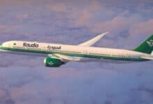 A Saudia airline aeroplane flying above the clouds during sunset, featuring a white fuselage with green and blue stripes, a green tail with a palm tree and two swords logo, and Arabic and English text on the side.