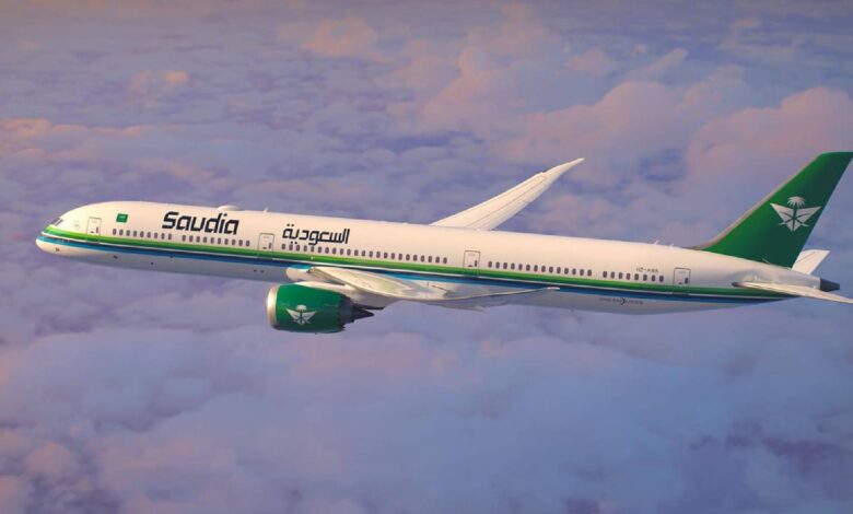 A Saudia airline aeroplane flying above the clouds during sunset, featuring a white fuselage with green and blue stripes, a green tail with a palm tree and two swords logo, and Arabic and English text on the side.