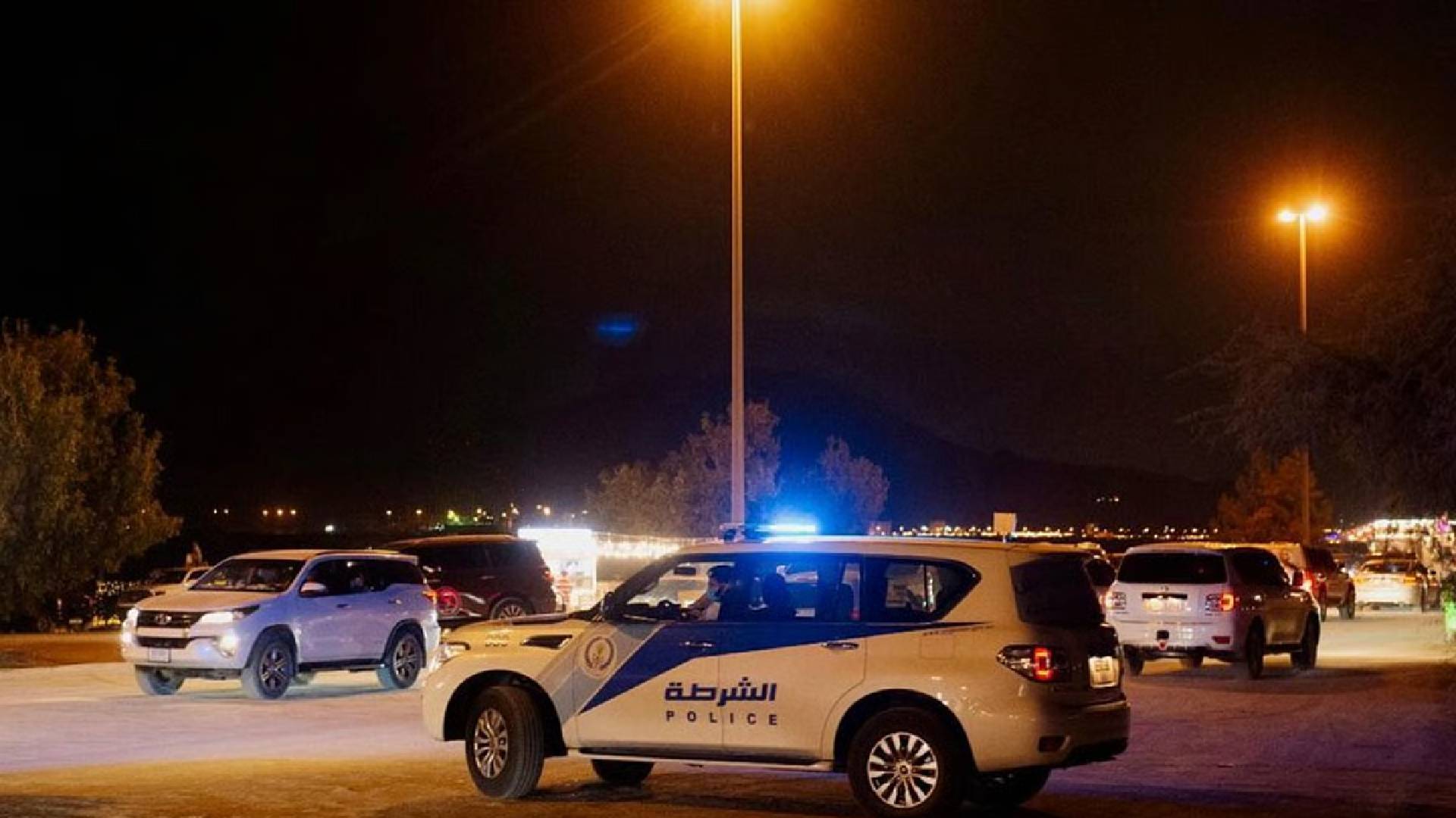 The image of a Sharjah Police patrol vehicle with flashing lights parked at night, surrounded by other cars under streetlights.