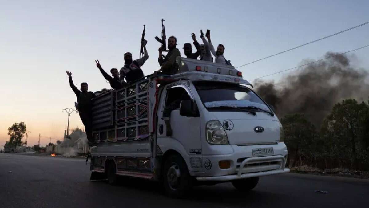A group of armed Bedouin fighters raise their weapons while riding on a white pickup truck along a street in Sweida province, with black smoke rising in the background during ongoing sectarian clashes.