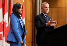 Canadian Prime Minister Mark Carney speaks at a podium during a press conference, accompanied by a senior official, with Canadian flags in the background.
