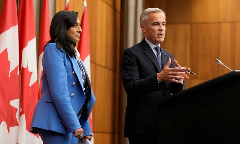 Canadian Prime Minister Mark Carney speaks at a podium during a press conference, accompanied by a senior official, with Canadian flags in the background.