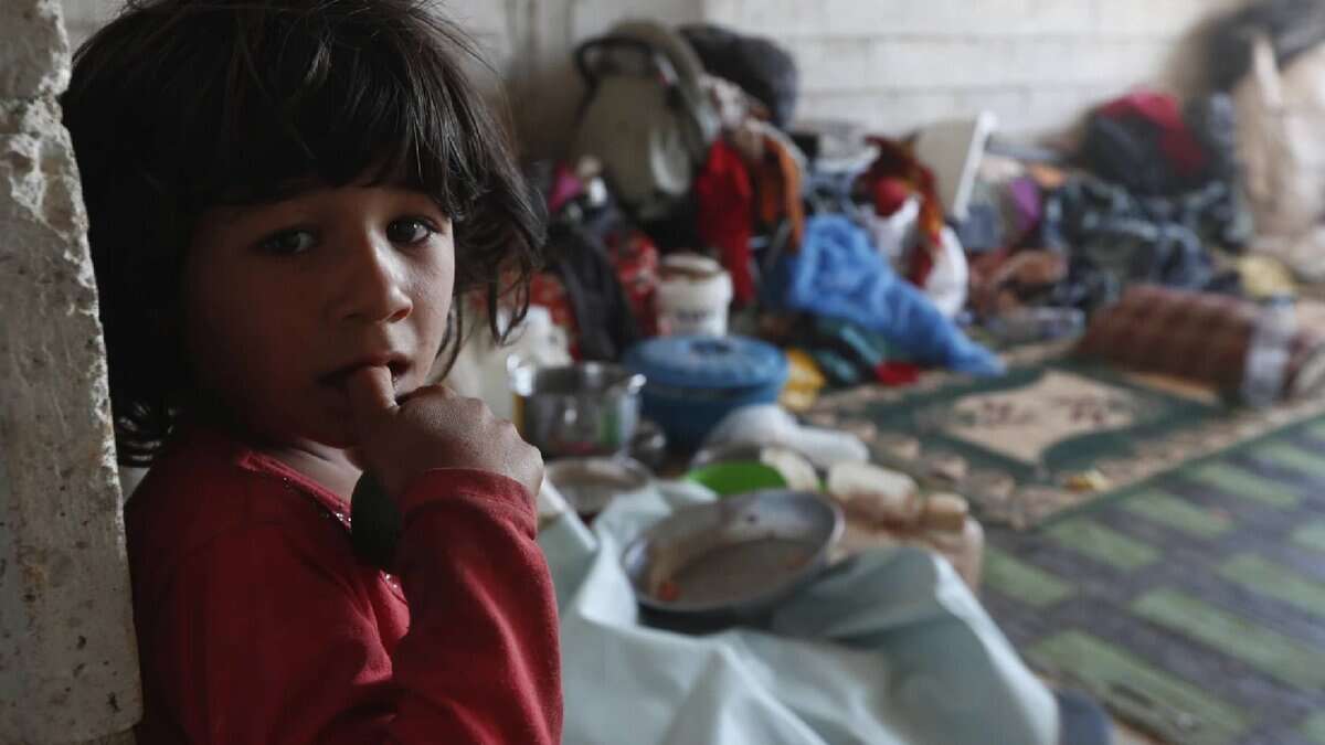 A displaced Bedouin girl in a red shirt stands inside a bare shelter, looking at the camera, with scattered household items and bedding behind her.