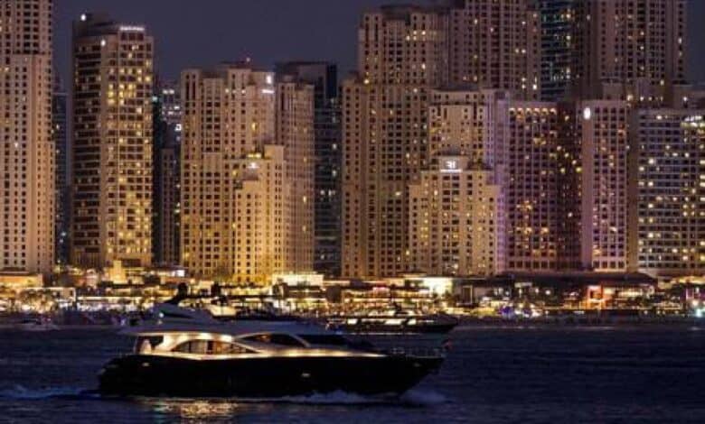 The image of a luxury yacht cruises past Dubai Marina skyscrapers illuminated at night, showcasing the city's vibrant waterfront lifestyle.