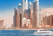 A luxury yacht cruises across the blue waters in front of Dubai Marina’s modern skyscrapers, under a clear sky.