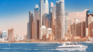 A luxury yacht cruises across the blue waters in front of Dubai Marina’s modern skyscrapers, under a clear sky.