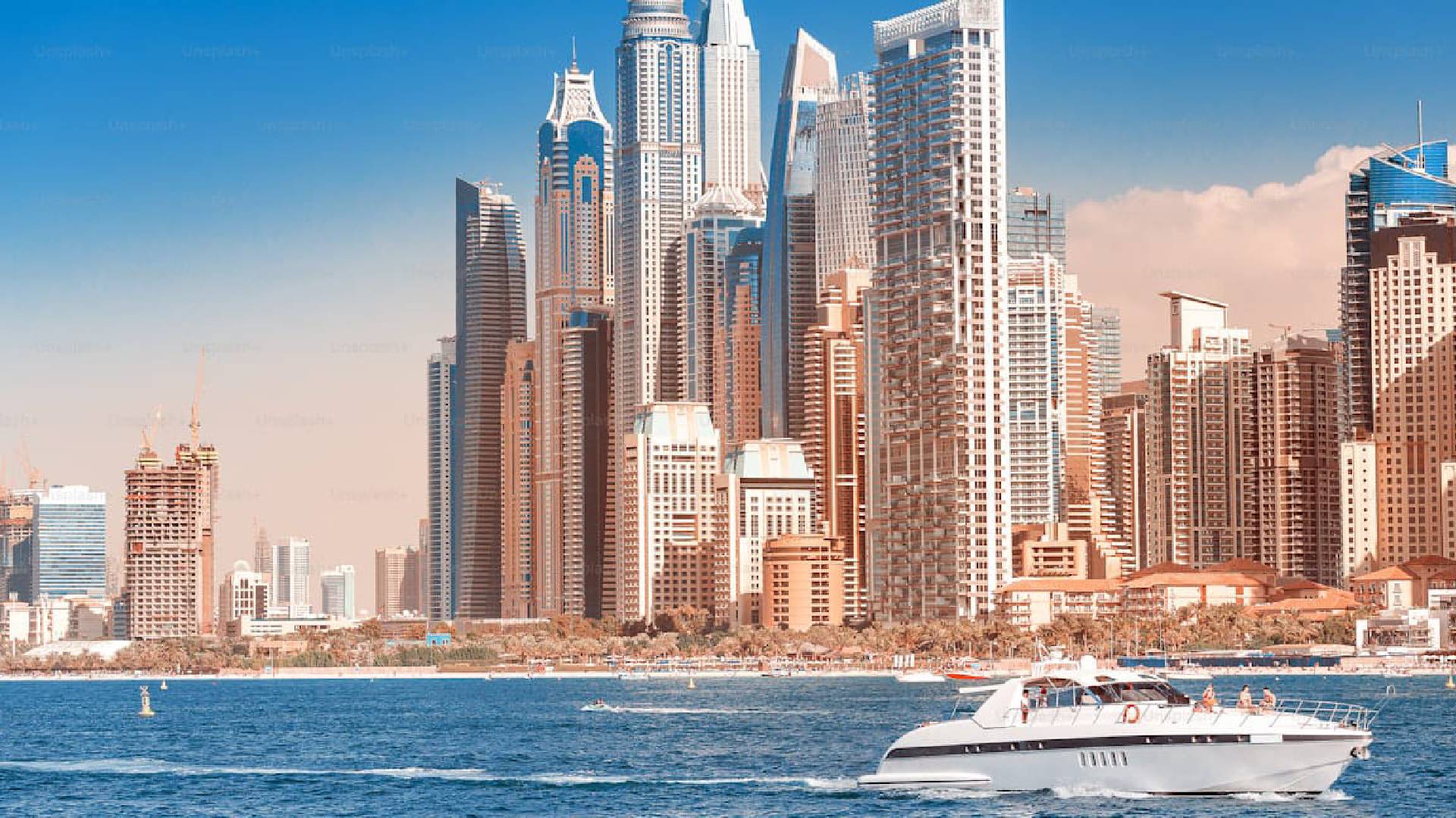 A luxury yacht cruises across the blue waters in front of Dubai Marina’s modern skyscrapers, under a clear sky.