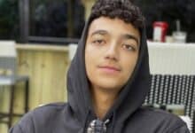 Edan Alexander, wearing a black hoodie and sunglasses around his neck, sits outdoors at a cafe.
