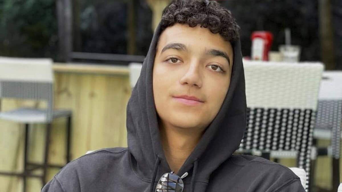 Edan Alexander, wearing a black hoodie and sunglasses around his neck, sits outdoors at a cafe.