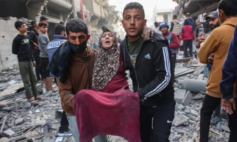 Two men carry an injured elderly woman through the rubble of destroyed buildings in Gaza, as others look on amidst the devastation.