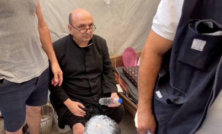 A priest with a bandaged leg sits on a bed inside a tent, surrounded by people offering assistance after an attack on Gaza's Holy Family Church.