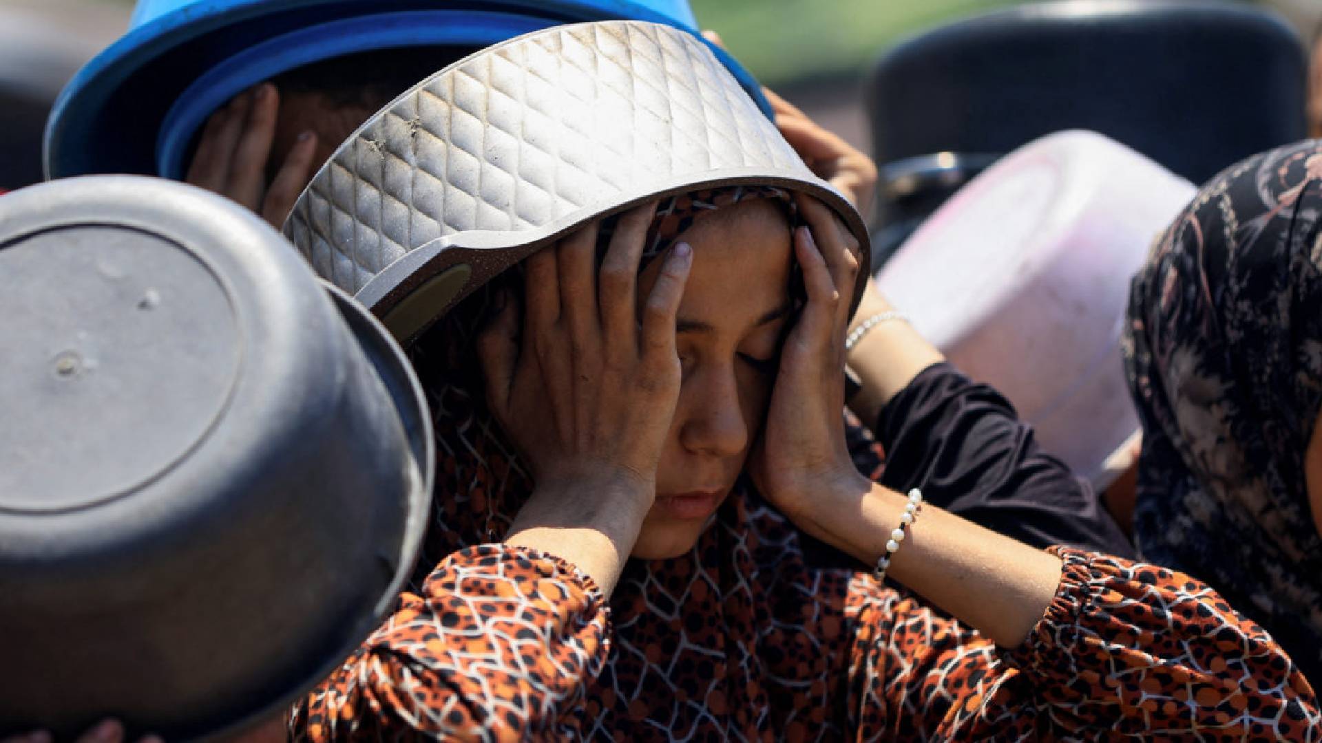 A Palestinian girl, with a cooking pot on her head for shade, rests her face in her hands while waiting in a long queue to receive food in Gaza City.
