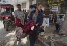 Two Palestinian men carry a body wrapped in blankets into Gaza City’s Shifa Hospital following an Israeli airstrike, as others look on outside the building.