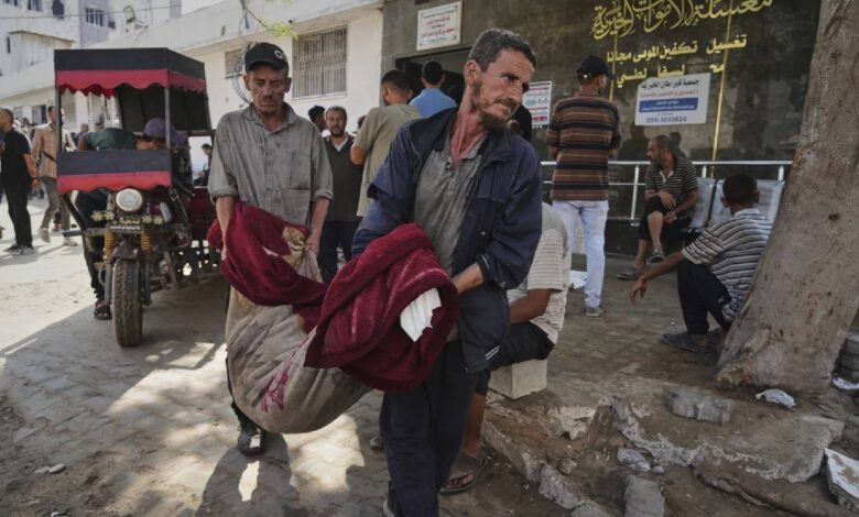 Two Palestinian men carry a body wrapped in blankets into Gaza City’s Shifa Hospital following an Israeli airstrike, as others look on outside the building.