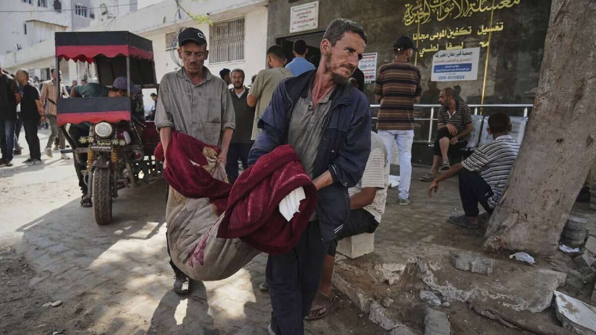 Two Palestinian men carry a body wrapped in blankets into Gaza City’s Shifa Hospital following an Israeli airstrike, as others look on outside the building.