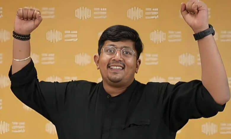 A joyful man with raised arms, wearing a black shirt and glasses, standing against a yellow patterned background.