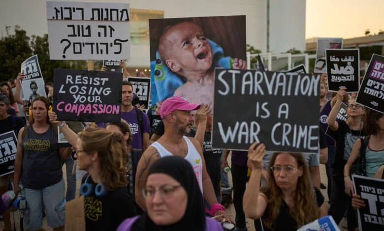 Israeli activists protest in Tel Aviv against the Gaza war, food restrictions, and forced displacement of Palestinians. Demonstrators hold signs reading “Starvation is a war crime” and “Resist losing your compassion,” alongside images of malnourished children.