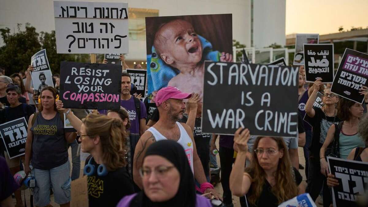 Israeli activists protest in Tel Aviv against the Gaza war, food restrictions, and forced displacement of Palestinians. Demonstrators hold signs reading “Starvation is a war crime” and “Resist losing your compassion,” alongside images of malnourished children.