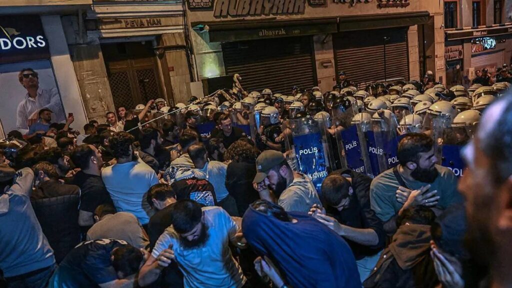 Riot police in helmets and shields confront a large crowd of protesters during a night-time clash in Istanbul.