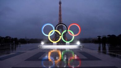 Olympic rings displayed on Trocadero Esplanade with Eiffel Tower in the background, wet pavement reflecting the rings under a rainy sky.