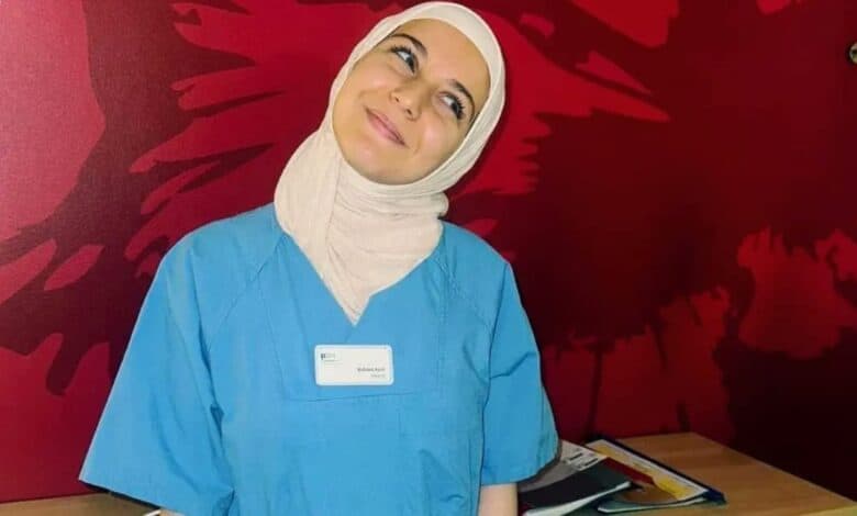 Smiling young woman in a blue nursing uniform and beige headscarf stands in front of a red wall with abstract patterns. She has a name badge that reads "Rahma Ayat".