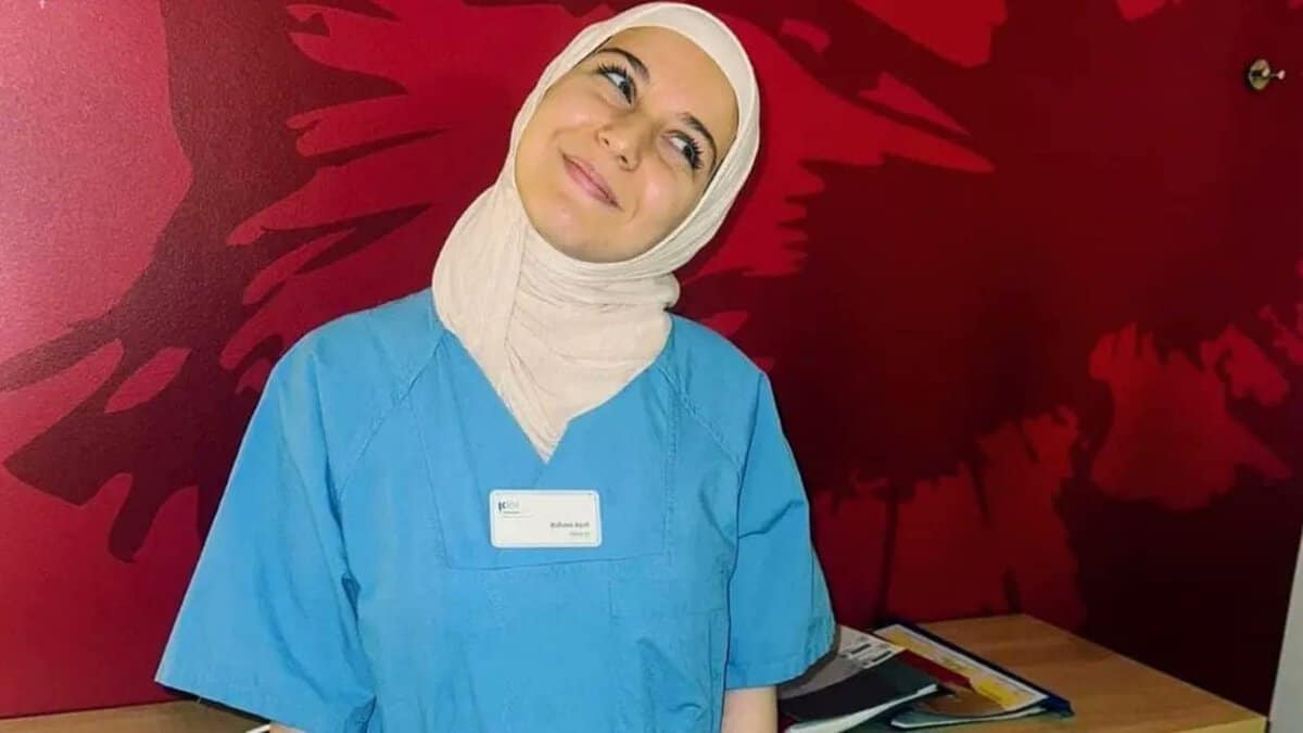 Smiling young woman in a blue nursing uniform and beige headscarf stands in front of a red wall with abstract patterns. She has a name badge that reads 