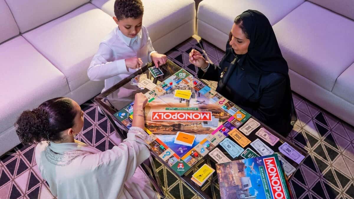 The image of a thrree people, including a child and two women, play the newly launched Riyadh edition of Monopoly at a modern indoor setting.