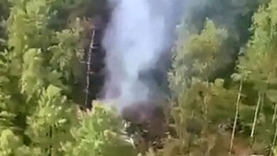 A plume of white smoke rises from a forested crash site surrounded by dense green trees in a mountainous area. The wreckage is partially visible among the trees.