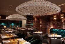 Luxurious interior of a Saudi café featuring elegant crystal chandeliers, plush seating, rich wood and patterned wall decor, with teal and gold accents. Tables are neatly set with amber glassware and napkins, creating a warm and upscale dining ambiance.