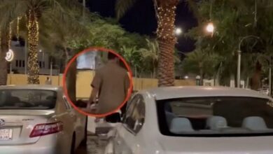 A man circled in red is seen walking between parked cars at night in a palm-lined street, reportedly wearing a night thobe. The scene is dimly lit with streetlights and decorative tree lights.