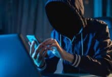 The image of a person in a dark hoodie with a hidden face sits at a desk, using a smartphone and a laptop in a dimly lit room with blue lighting, suggesting a secretive or cyber activity.
