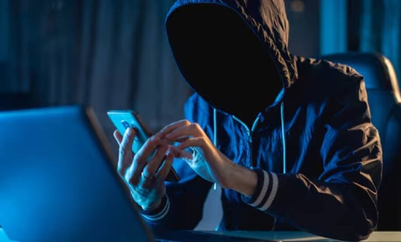 The image of a person in a dark hoodie with a hidden face sits at a desk, using a smartphone and a laptop in a dimly lit room with blue lighting, suggesting a secretive or cyber activity.